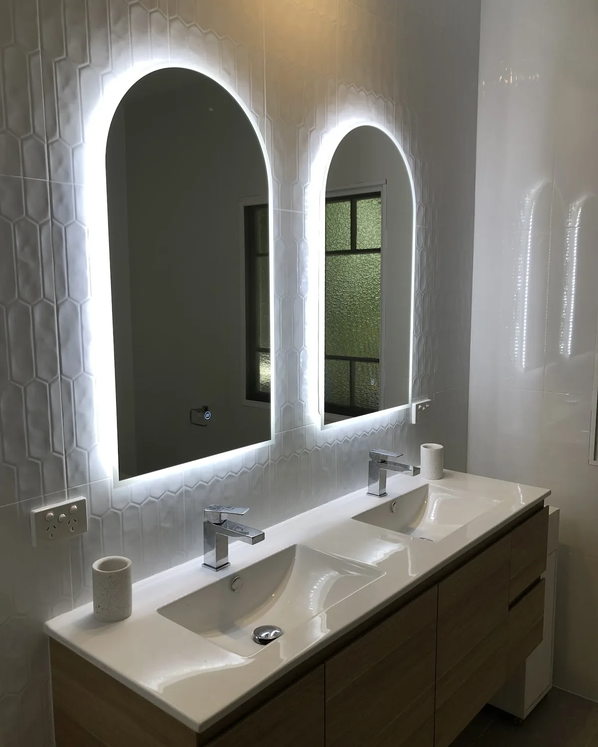 Two backlit arched mirrors above a double vanity in a white tiled bathroom