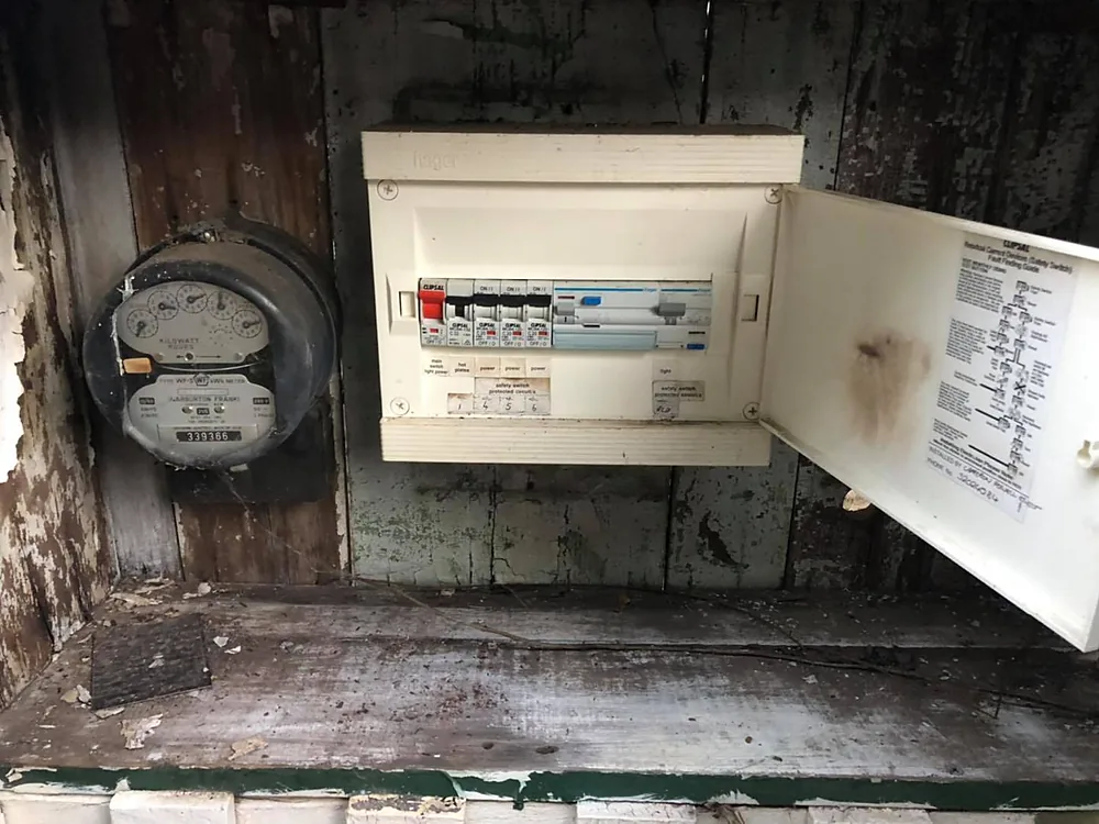 An old meter board with a ceramic meter and ageing fuses on a weathered wall, before an Aird's upgrade in Ipswich